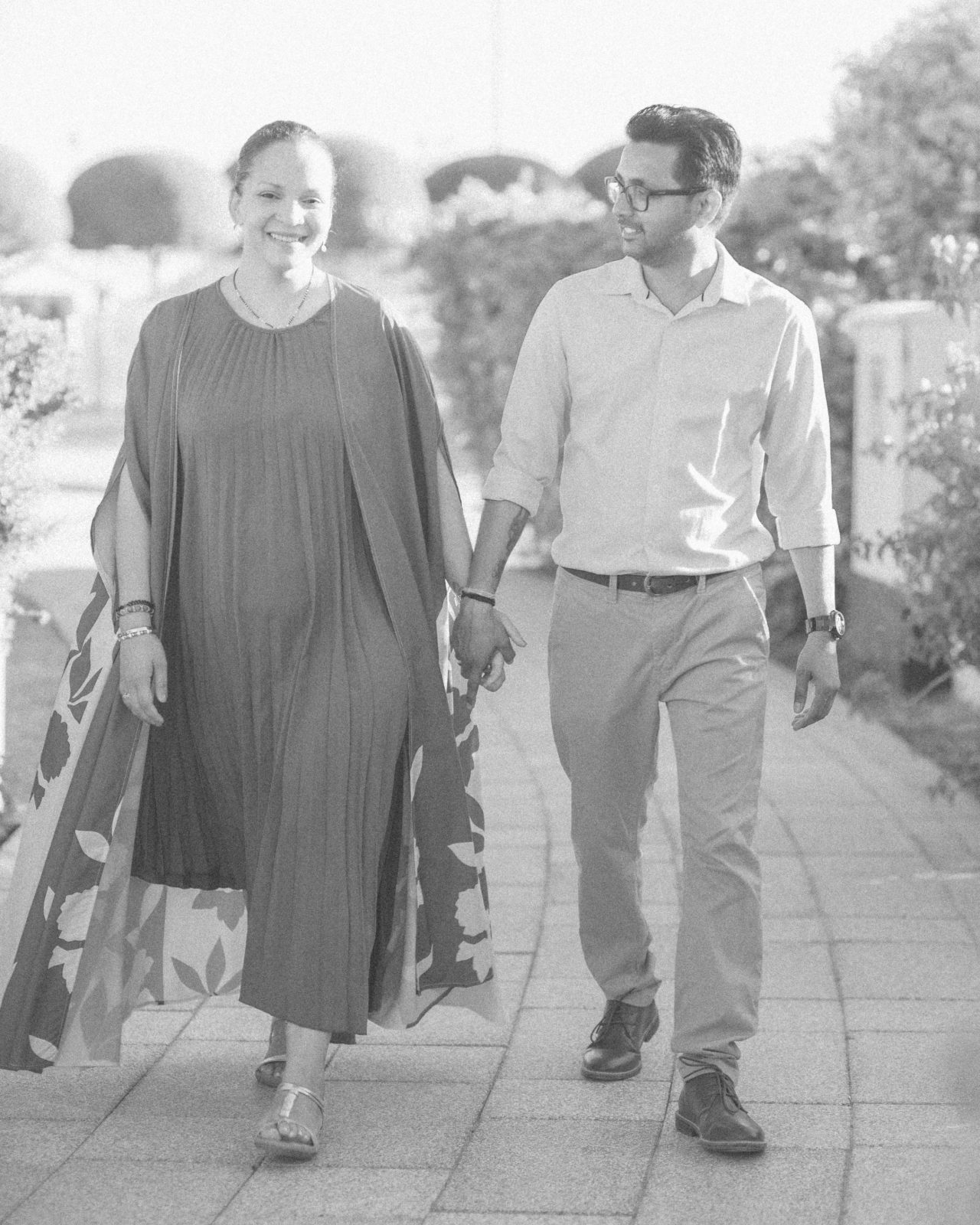 Black and white photo of a couple holding hands and walking together through a garden at golden hour