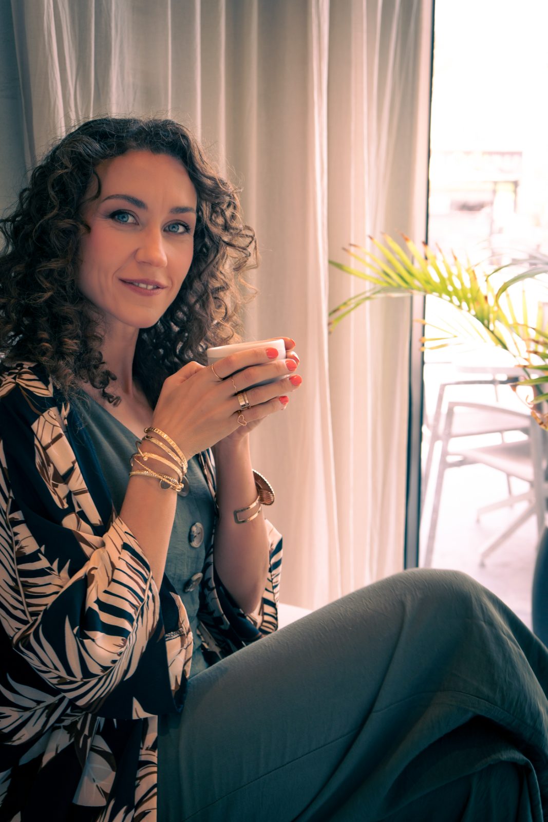 Individual portrait of a woman with curly hair smiling softly while holding a cup in warm window light
